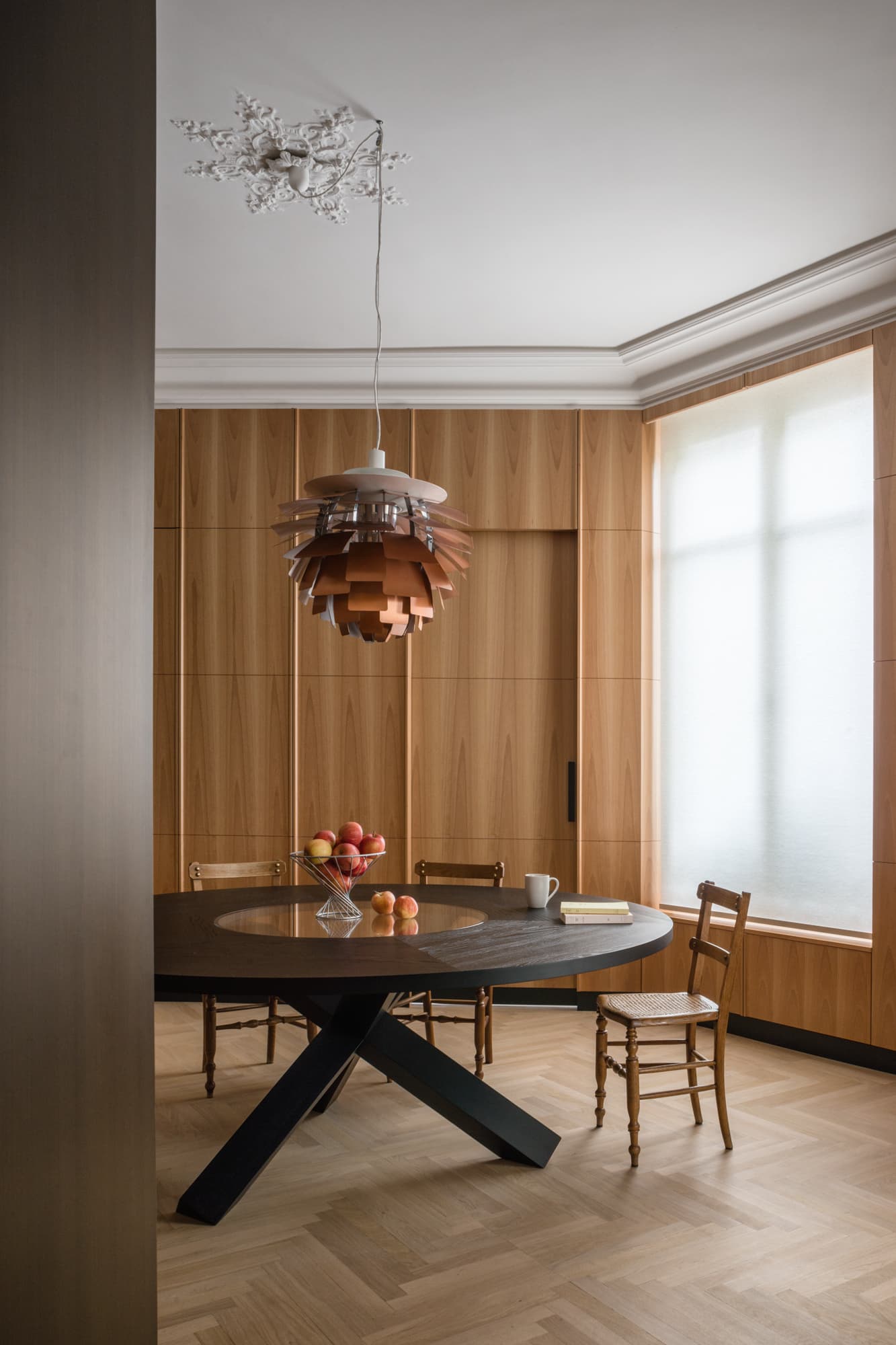 Vue de la salle à manger avec table ronde en bois teinté sombre, suspension PH Artichoke en cuivre, chaises anciennes dépareillées, boiseries en placage de bois clair et parquet en chevrons.