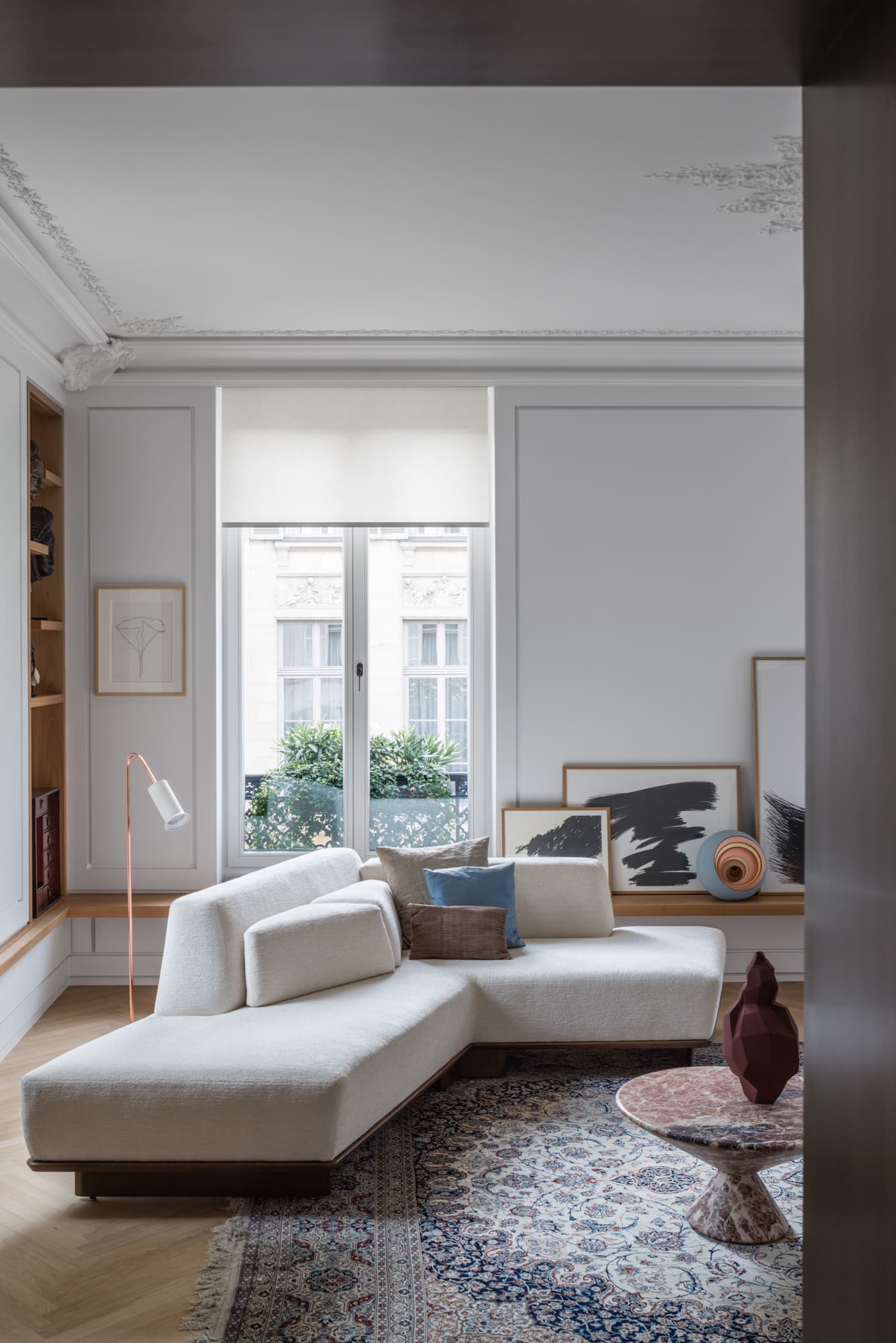 Vue du séjour avec canapé modulable en tissu écru, boiseries et moulures d'origine, bibliothèque intégrée présentant une collection d'œuvres d'art, tapis ancien et grande fenêtre à la française.