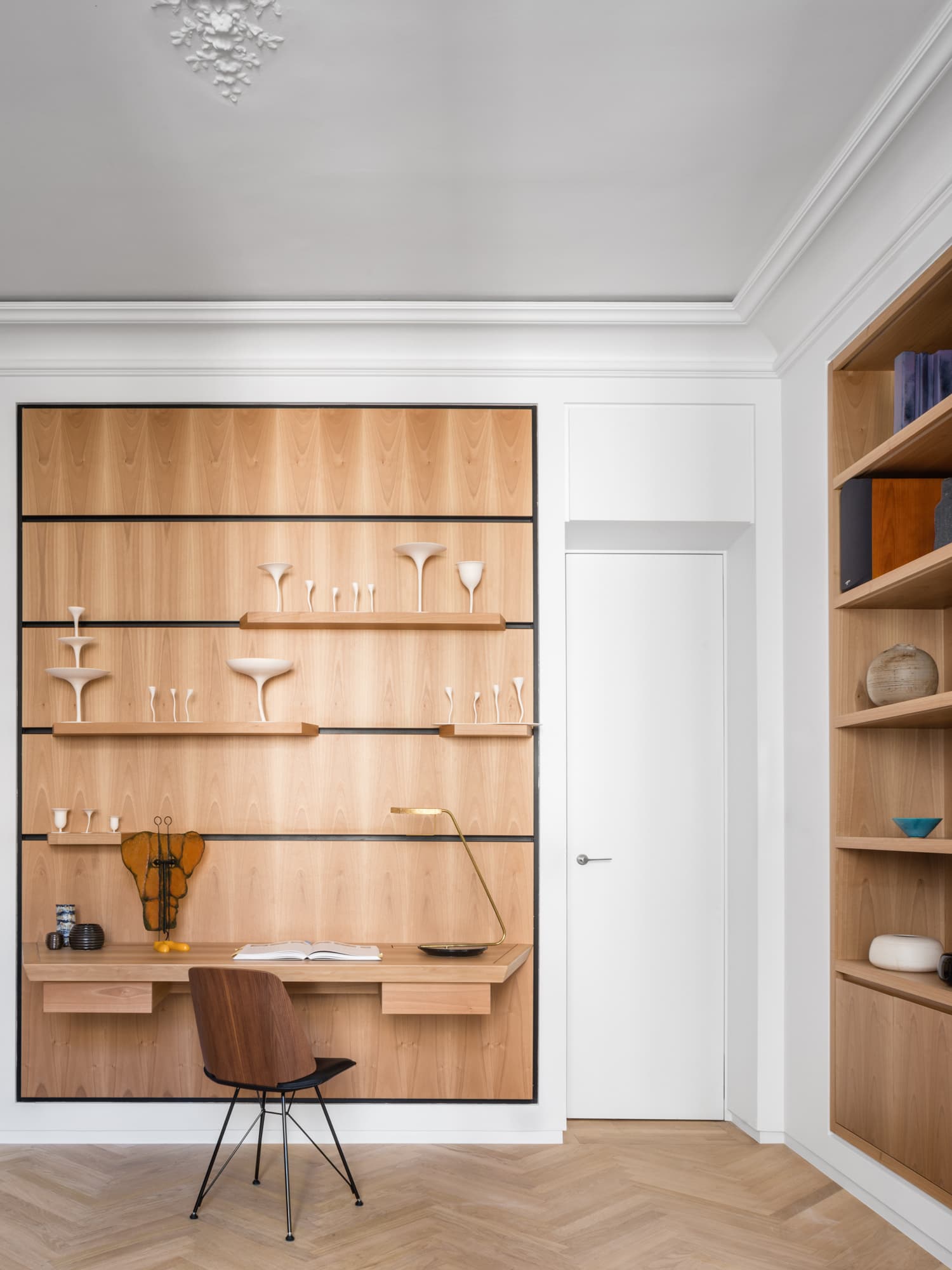Vue du bureau intégré sur mesure en bois clair, avec étagères présentant une collection de céramiques et d'objets d'art, lampe en laiton et chaise cannée.