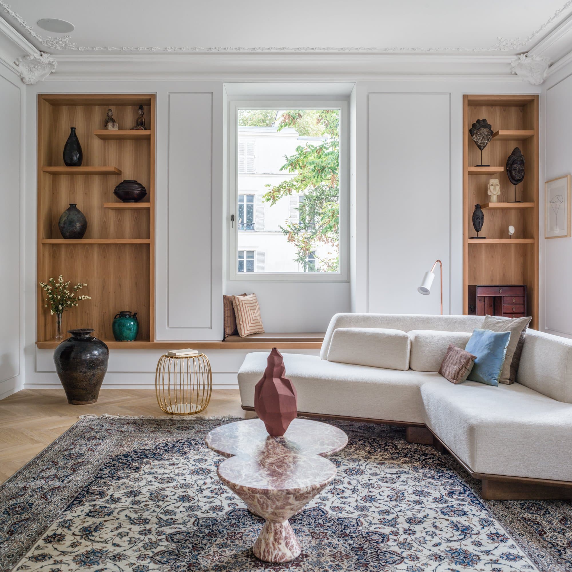 Vue du séjour avec bibliothèques sur mesure encadrant la fenêtre, présentant une collection de céramiques et de masques africains, canapé écru, tapis ancien et table basse en marbre.