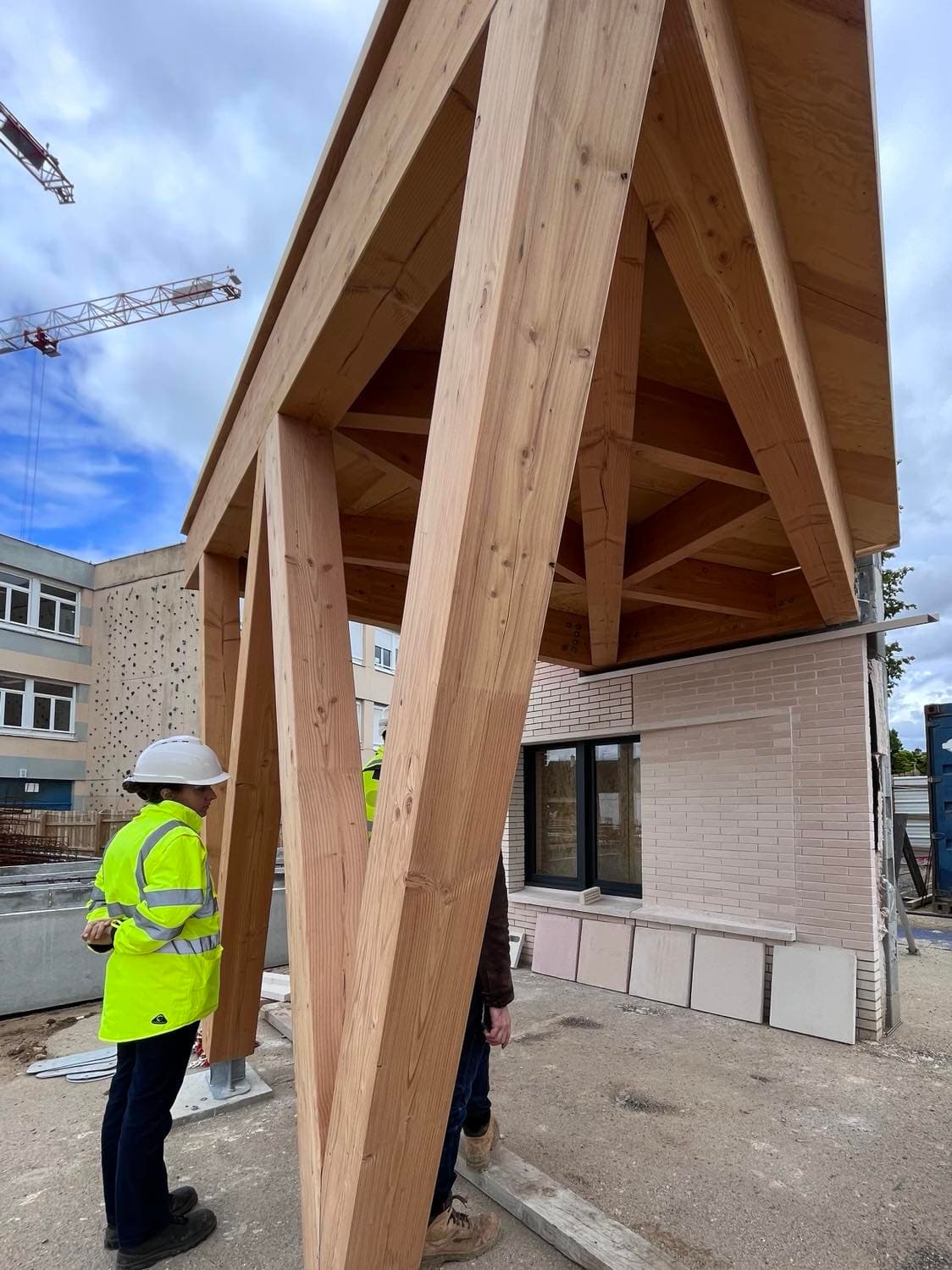 Photo de chantier de la charpente bois de l'auvent d'entrée en cours de montage, avec ouvriers casqués et grue en arrière-plan.